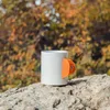 Weißer Kaffeebecher mit orangefarbenem Klettergriff; platziert auf einem Felsen in der Natur. Náhľad obrázku produktu