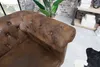 A close-up view of a brown Chesterfield armchair with button tufting, showing the armrest and part of the seat. The background includes a brick wall, a plant, and a shelf with white boxes. Náhled obrázku produktu