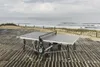 A ping pong table is set up on a wooden deck near a beach with the ocean in the background. Produktbild-Vorschau
