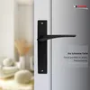 A black door handle with a long plate is mounted on a white door. The background is blurred, showing a room with furniture and decorative items. Produktbild-Vorschau
