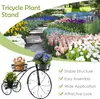 A black metal tricycle-shaped plant stand with three baskets holding potted plants, set against a backdrop of a colorful flower garden. Produktbild-Vorschau