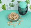 A bowl of mini wooden clothespins sits on a teal placemat next to a teal candle in a glass holder, with a tropical leaf patterned wallpaper in the background. Náhled obrázku produktu