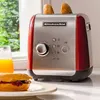 A red and silver KitchenAid toaster sits on a countertop next to a glass of orange juice and a breakfast sandwich. Produktbild-Vorschau