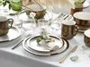 A beautifully set dining table with metallic bronze and white stoneware plates, mugs, and bowls, accented with greenery and candles. Náhľad obrázku produktu