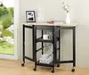 A folding kitchen cart with a wooden top and black metal frame, set up next to a window with a view of greenery. It is decorated with a framed picture, books, a coffee maker, a mug, and a wine bottle. Produktbild-Vorschau