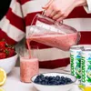 A person is pouring a pink smoothie from a blender into a glass, with ingredients like strawberries, blueberries, and cans of coconut water visible. Produktbild-Vorschau