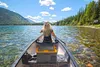 A person is paddling a canoe on a lake with mountains and trees in the background. A cooler is placed in the canoe. Produktbild-Vorschau