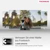 Three televisions are displayed in front of a blurred background of the Eiffel Tower and a park scene. The main television in the foreground shows a woman carrying a child on her back. Text and a warranty badge are present below the televisions. Produktbild-Vorschau