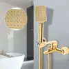 A gold-colored shower faucet and hand shower head are mounted on a gray wall, with a blurred white bathtub in the background. Podgląd zdjęcia produktu