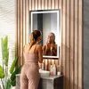 A woman is applying makeup in front of a lighted mirror in a modern bathroom. Produktbild-Vorschau