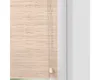 A close-up view of a natural bamboo roller blind hanging next to a white wall. Náhled obrázku produktu