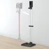 A floor stand for a cordless vacuum cleaner is shown against a white wall and wooden floor. A faded image of a vacuum cleaner is shown on the left, with an arrow pointing towards the stand, indicating it is a storage solution. Produktbild-Vorschau