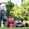 A person is using a red and black lawnmower on a green lawn with trees in the background. Produktbild-Vorschau
