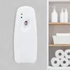 A white automatic air freshener dispenser mounted on a tiled wall, with a shelf containing soap dispensers and toilet paper rolls visible in the background. Produktbild-Vorschau