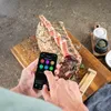 A person is using a smartphone to control a meat thermometer inserted into a large cut of raw meat on a wooden cutting board. Náhled obrázku produktu