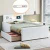 A white upholstered bed with a headboard featuring storage compartments and a built-in light. The bed has a pull-out drawer underneath, and is shown with pillows and bedding. Produktbild-Vorschau