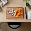 A wooden cutting board with sliced carrots, cucumbers, and red onions on a wooden table, next to a knife and a folded white cloth. Produktbild-Vorschau
