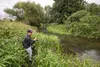 A person is fishing in a narrow river surrounded by lush green vegetation. Produktbild-Vorschau
