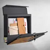 A dark gray mailbox with a wood-look flap is mounted on a textured light gray wall. The mailbox is open, revealing letters and envelopes inside. A newspaper is tucked into a compartment at the bottom. Produktbild-Vorschau