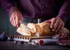 A person is slicing a baguette with a bread knife on a cutting board, with various accompaniments like olives, figs, and cured meat visible. Voir les images du produit