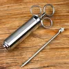 A stainless steel meat injector and its needle are placed on a wooden surface. Voir les images du produit