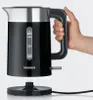 A hand holding a black electric kettle with a silver top and a water level indicator. The kettle is shown above its power base. Produktbild-Vorschau