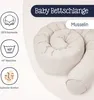 A coiled baby bed snake made of muslin fabric, with icons indicating material, washability, and comfort. Produktbild-Vorschau