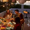 People enjoying a meal outdoors at night, with string lights and a patio heater. Anteprima dell’immagine prodotto