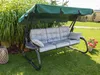A garden swing with cushions is set up on a lawn, with tall green plants in the background and a pot of pink flowers to the side. Podgląd zdjęcia produktu