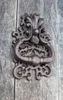 A close-up of an ornate, dark brown cast iron door knocker mounted on a weathered wooden door. Produktbild-Vorschau