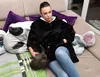 A woman sits on a couch wearing a black hooded blanket, holding a cat. Produktbild-Vorschau