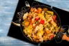 A close-up overhead view of a pan filled with cooked vegetables, placed on a black surface with a floral design, which is on a blue wooden table. Náhľad obrázku produktu