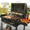 A Royal Gourmet charcoal grill is set up outdoors with food cooking on it, and various items like fruits, sauces, and a cutting board are placed around it. Produktbild-Vorschau