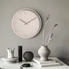 A round clock with embossed numbers hangs on a gray wall above a table with decorative vases and a stone. Anteprima dell’immagine prodotto