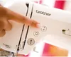 A close-up shot of a person's finger pressing a button on a white Brother sewing machine. Produktbild-Vorschau