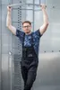 A man wearing overalls and a t-shirt stands in front of a metal structure, holding onto a metal ring above his head. Náhled obrázku produktu