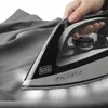 A hand is holding a Black+Decker iron, pressing it onto a gray fabric. The iron is illuminated from the bottom, highlighting its use. Produktbild-Vorschau