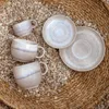 Three coffee cups and two saucers are arranged on a woven mat covered with small stones. Anteprima dell’immagine prodotto