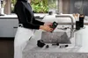 A person is washing a coffee machine part under a faucet in a modern kitchen. Voir les images du produit