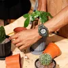 A person's arm wearing a green digital watch is shown among several potted plants and empty pots. Produktbild-Vorschau