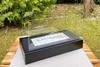 A black freestanding bio-fireplace with a glass wind guard sits on a table outdoors, with greenery in the background. Produktbild-Vorschau