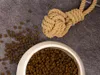 A bowl of dog food and a rope toy are shown on a gray surface. Produktbild-Vorschau