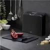A kitchen counter scene featuring a set of black cutting boards with colored tabs, a pomegranate, a knife, and kitchen utensils hanging in the background. Anteprima dell’immagine prodotto