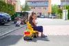 A young girl is sitting on a yellow toy trailer with a crane attachment, smiling at the camera. Another child is visible in the background, and houses and cars are in the distance. Náhled obrázku produktu
