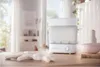 A white baby bottle sterilizer is placed on a wooden table in a bright, airy room with a blurred background of a living space. Produktbild-Vorschau