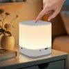 A hand is shown touching a white LED nightstand lamp on a bedside table next to a book and a vase with flowers. Voir les images du produit