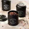 Three black metal canisters for storing tea, coffee, and sugar, with a lid off one canister showing tea leaves. Produktbild-Vorschau