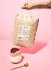 A hand holding a large bag of oats above a glass of yogurt with berries and a spoon. Produktbild-Vorschau