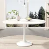 A white oval dining table with a pedestal base is shown in a well-lit room with a large window overlooking trees. Náhled obrázku produktu