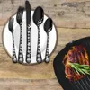 A set of black Christmas-themed cutlery is arranged on a white plate, with a grilled steak in a pan in the foreground. Podgląd zdjęcia produktu